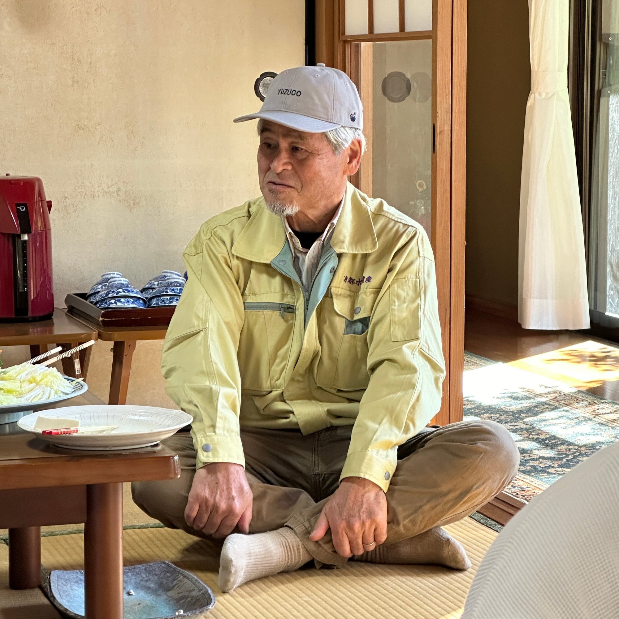 A man sitting cross legged wearing a YUZUCO hat